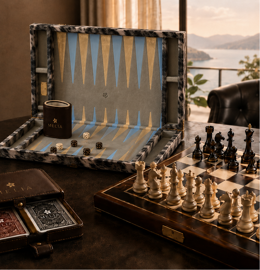 Promotional graphic for MELIA luxury games, featuring a backgammon board set, dice, a small branded cup, and a leather case on a dark table with large text reading 'Discover the World of Luxury Games' and 'MELIA'.