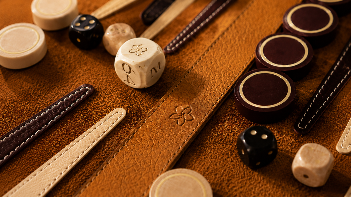 Close-up of a handcrafted MELIA leather backgammon set featuring the signature five-petal flower emblem, wooden checkers and dice.